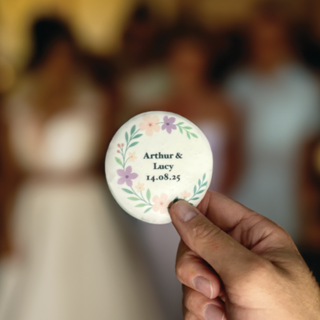 Hand holding a round biscuit with floral design and text in front of a blurred background