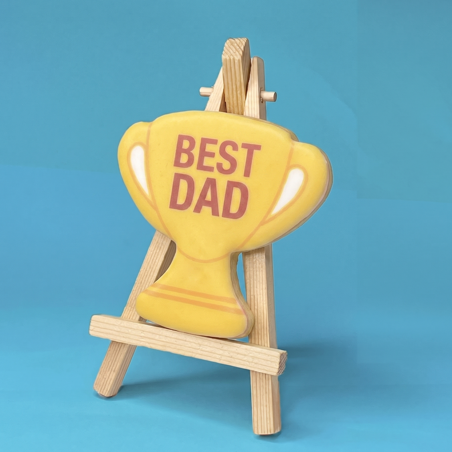 Yellow trophy-shaped biscuit with 'BEST DAD' text on a wooden stand against a blue background